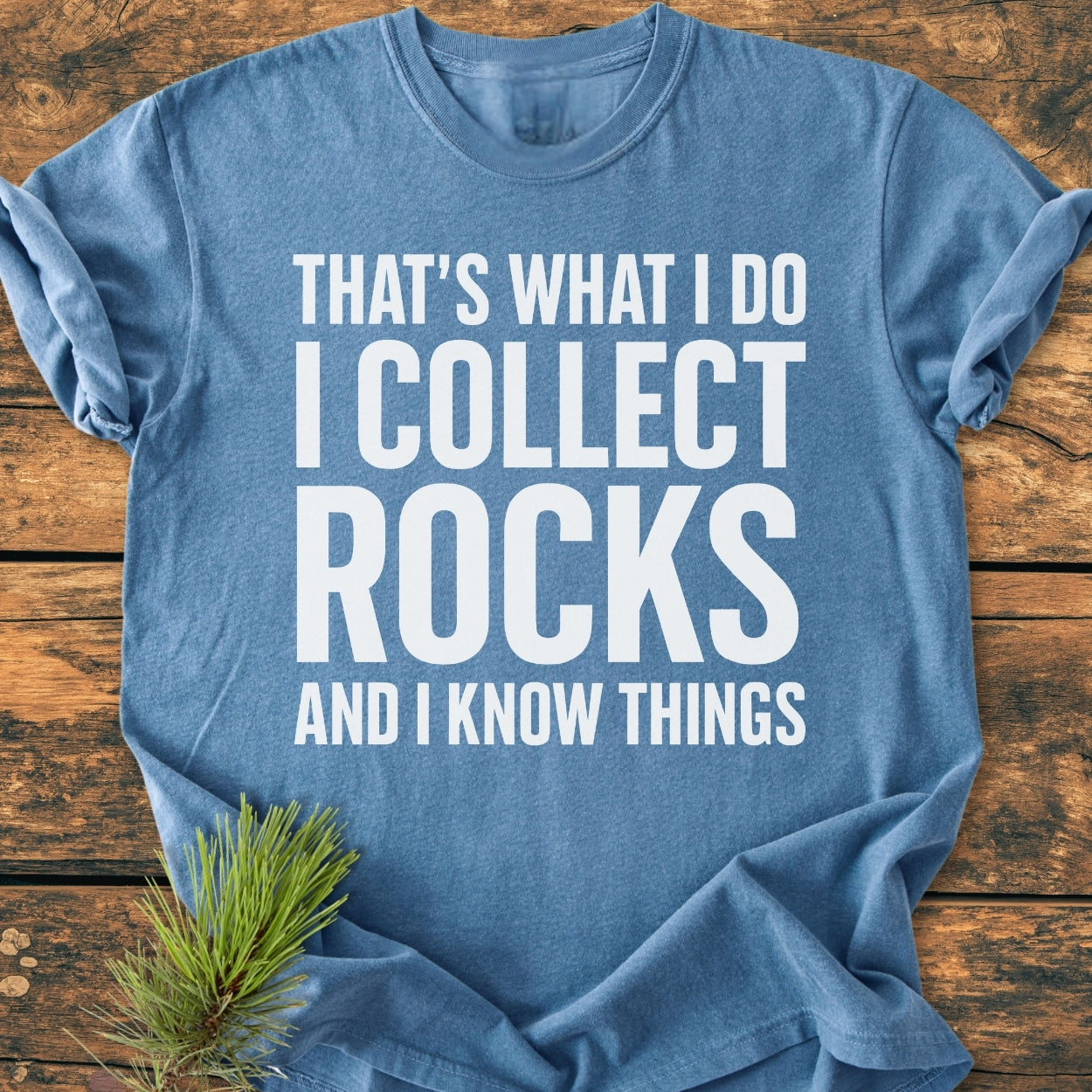 I Collect Rocks - Vintage Wash Tee: A blue short-sleeve T-shirt on a wooden surface with white block letters stating, “THAT’S WHAT I DO I COLLECT ROCKS AND I KNOW THINGS,” featuring a relaxed fit and durable stitching.