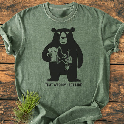 That Was My Last Hike - Vintage Wash Tee: A forest-green tee featuring a grumpy bear with beer and a cartoon hiker skeleton. Laid flat on wood, showcasing its relaxed fit and durable stitching.