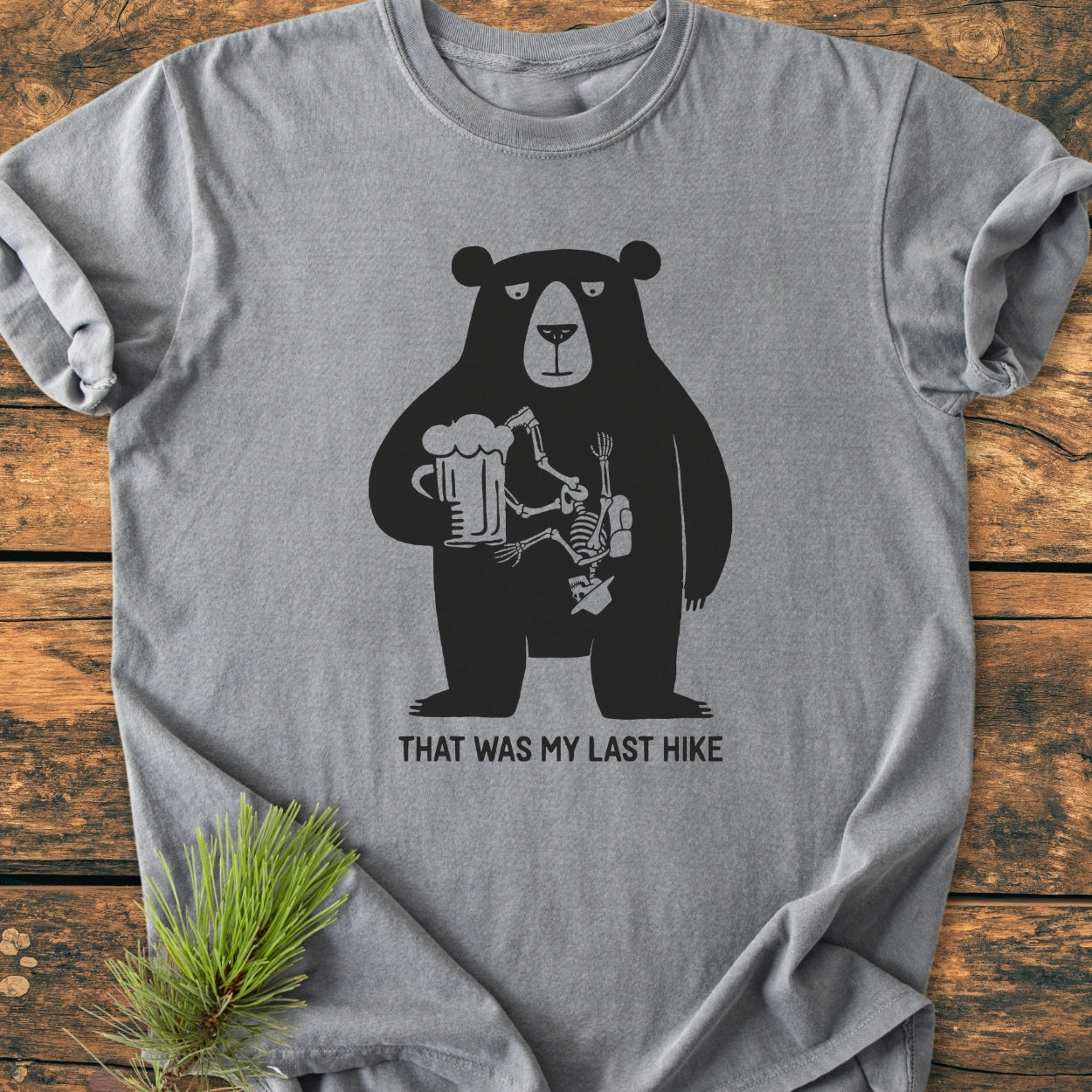 That Was My Last Hike - Vintage Wash Tee features a grumpy bear with a hiker's skeleton inside, holding a beer mug, captioned humorously, laid flat to display the unique design.