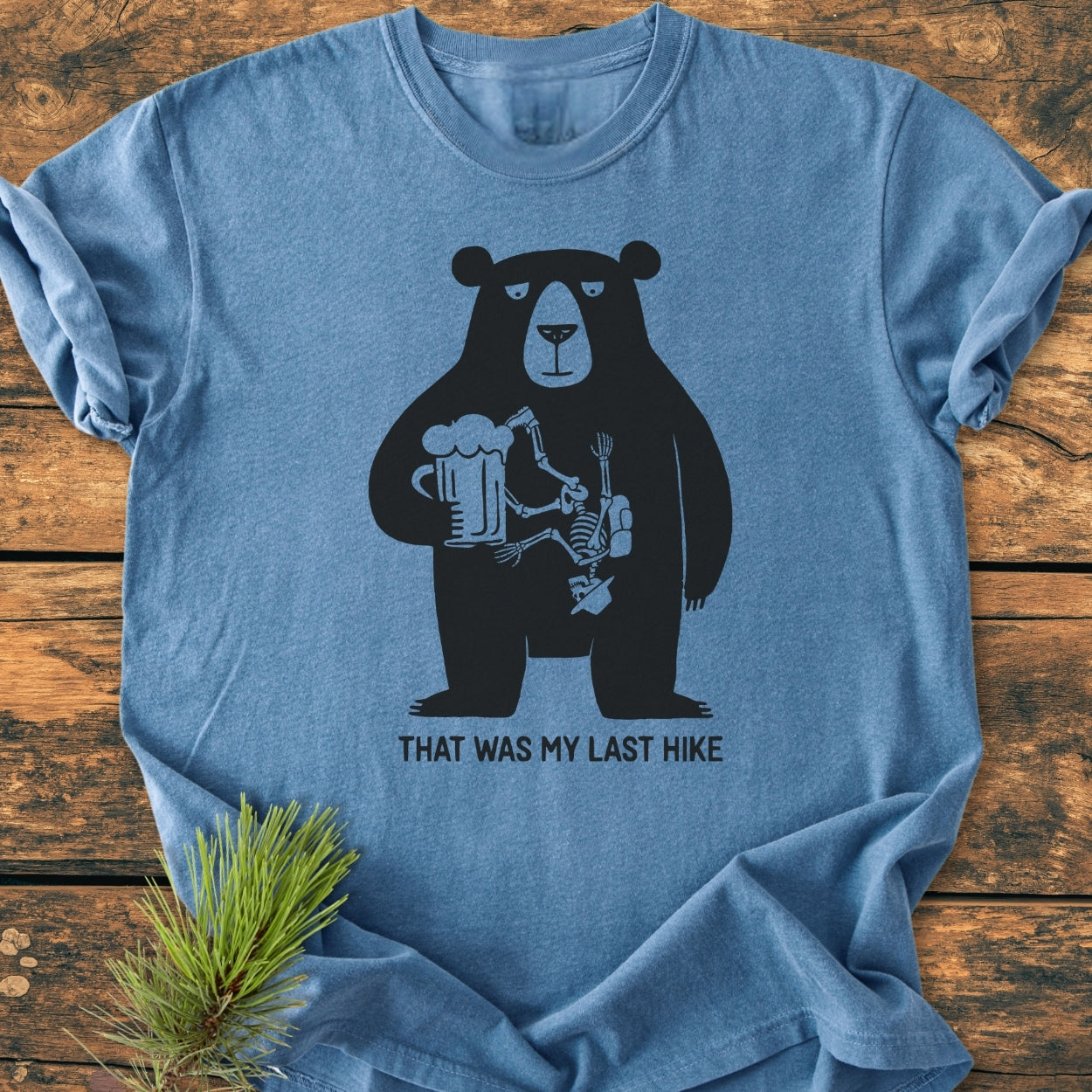 That Was My Last Hike - Vintage Wash Tee features a black bear with a beer mug and hiker skeleton graphic on a heather-blue backdrop, displayed on a rustic wooden surface.