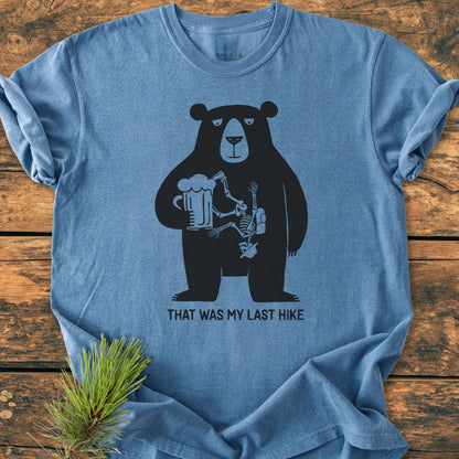 That Was My Last Hike - Vintage Wash Tee features a black bear with a beer mug and hiker skeleton graphic on a heather-blue backdrop, displayed on a rustic wooden surface.
