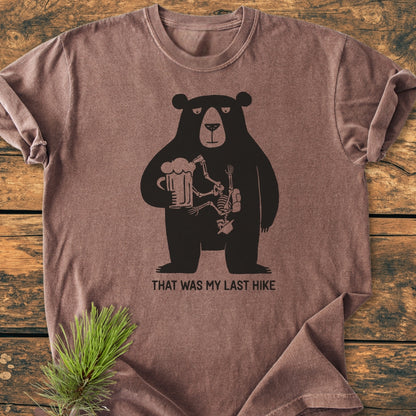 That Was My Last Hike - Vintage Wash Tee features a cartoon bear with a beer mug, revealing a hiker's skeleton inside, captioned humorously, laid on a wooden surface with a pine sprig.