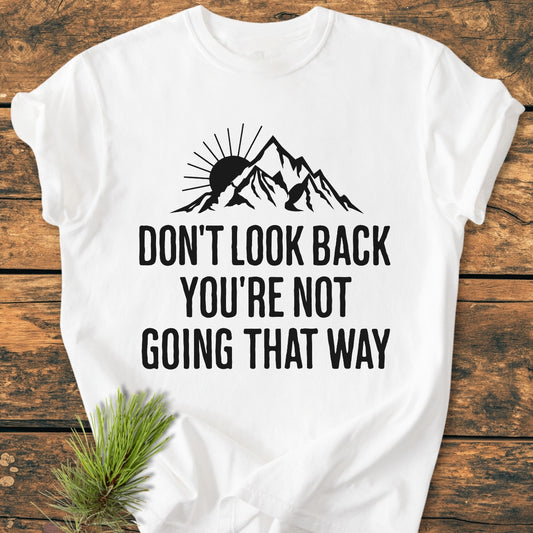 Don't Look Back - Vintage Wash Tee features a black stylized mountain range with a sun and motivational slogan on a white T-shirt, displayed on a wooden surface.