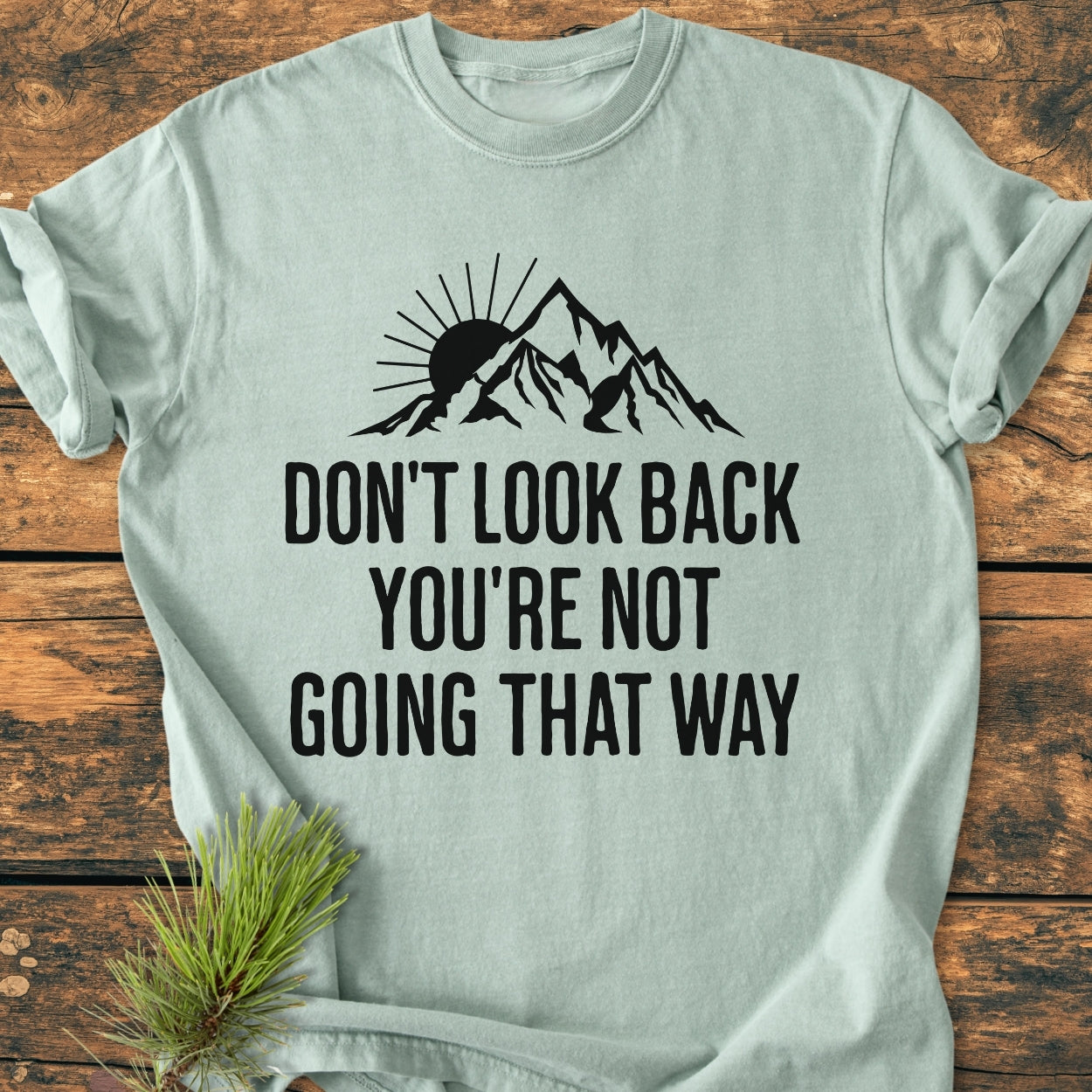 Don't Look Back - Vintage Wash Tee features a mountain range graphic with a sun, slogan beneath. Laid flat on a wooden surface, showcasing cozy, relaxed fit, 100% ring-spun cotton.