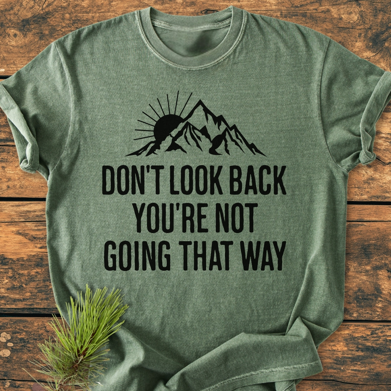Don't Look Back - Vintage Wash Tee features a black sun and mountain graphic with a motivational slogan on a heather-green, relaxed-fit, 100% cotton T-shirt, displayed on a wood surface with pine needles.