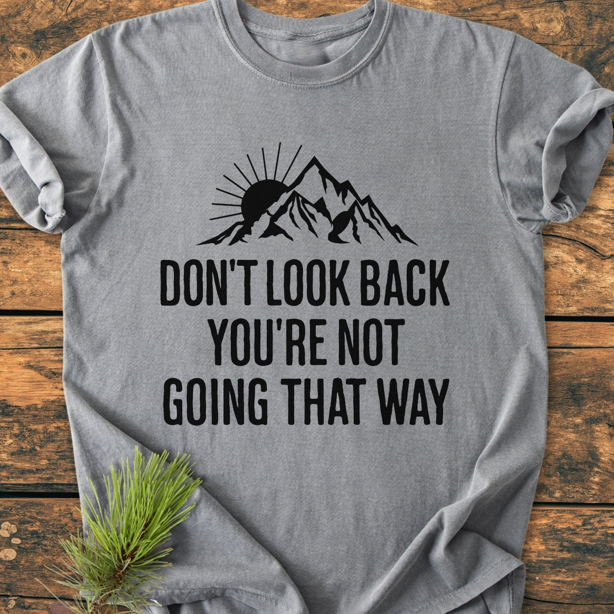 Don't Look Back - Vintage Wash Tee; a heather-gray shirt with a mountain silhouette and motivational motto, laid on wood with pine needles. Made of soft, garment-dyed cotton, offering comfort and durability.