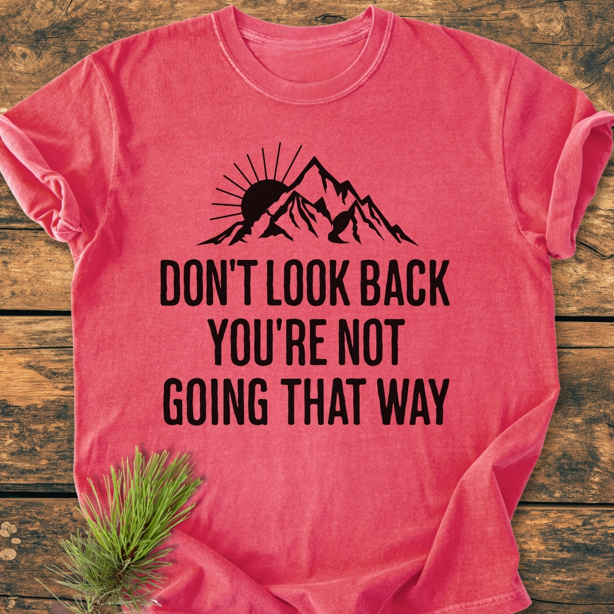 Don't Look Back - Vintage Wash Tee: Coral-red T-shirt with a stylized mountain and sun graphic, slogan in bold letters, laid flat on wood, with a pine sprig accent.