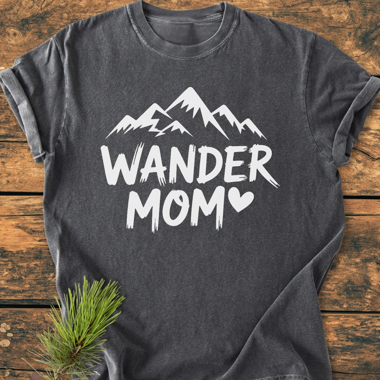 Wander Mom - Vintage Wash Tee features bold white 'WANDER MOM' text and mountain silhouette on a dark-gray T-shirt, laid flat on wood, with a pine sprig accentuating its rustic charm.