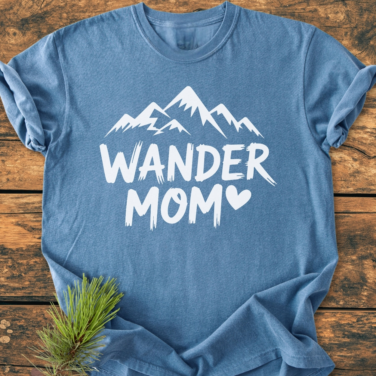 Heathered blue Wander Mom - Vintage Wash Tee with a stylized mountain range and heart-accented MOM, laid on rustic wood with a pine sprig, highlighting its cozy, relaxed fit.