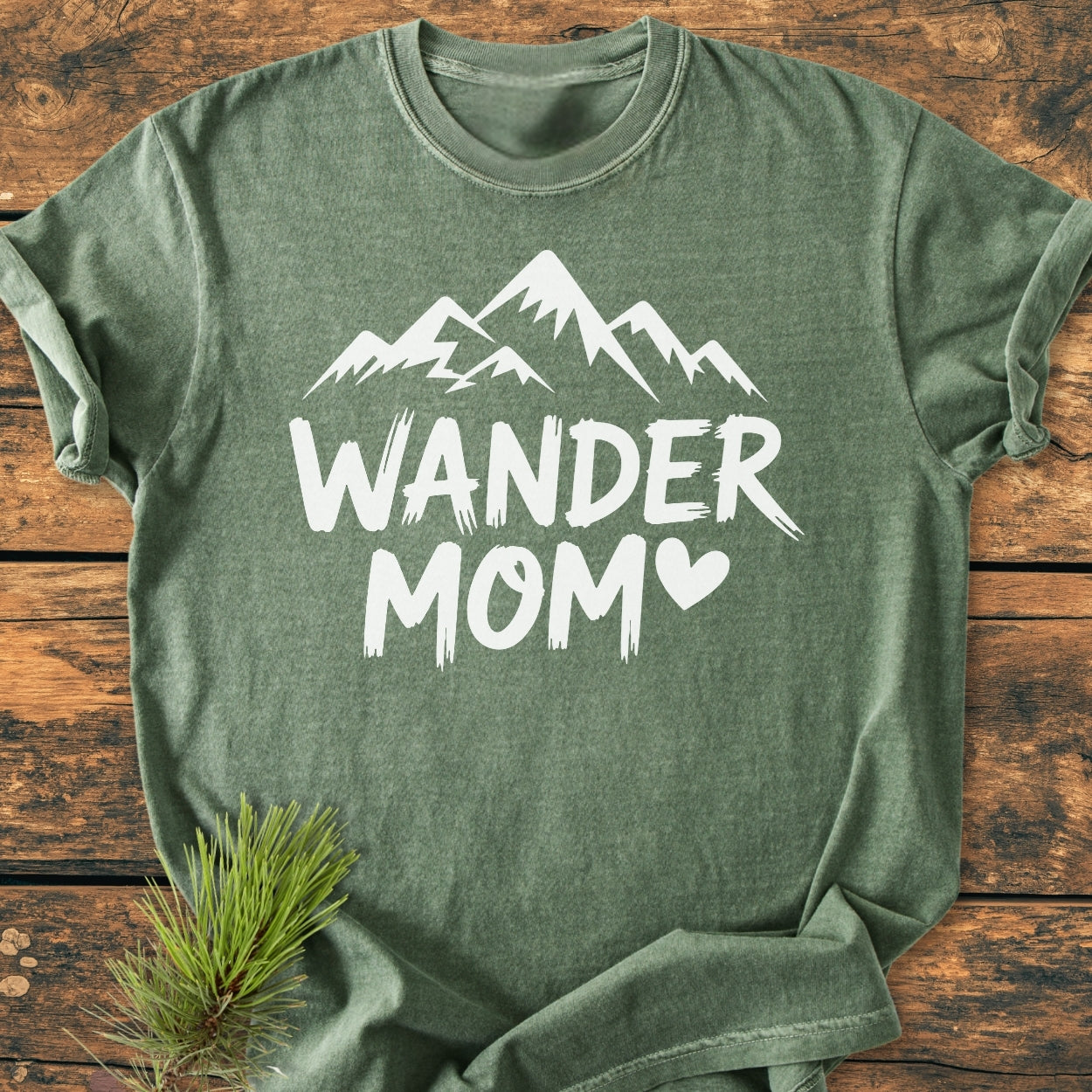 Heather-green Wander Mom - Vintage Wash Tee with a white stylized mountain and heart design, displayed flat on wood with a pine sprig, showcasing its relaxed fit and soft-washed texture.