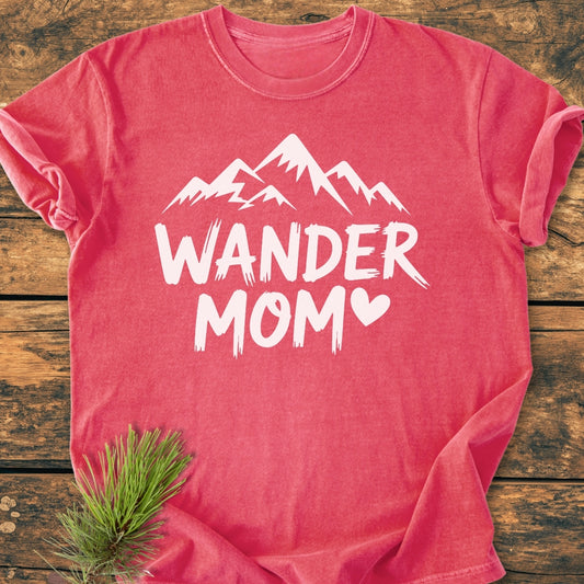 Wander Mom - Vintage Wash Tee, featuring a mountain silhouette and heart, folded on a wooden surface with a pine sprig, crafted from soft-washed, durable, ring-spun cotton.
