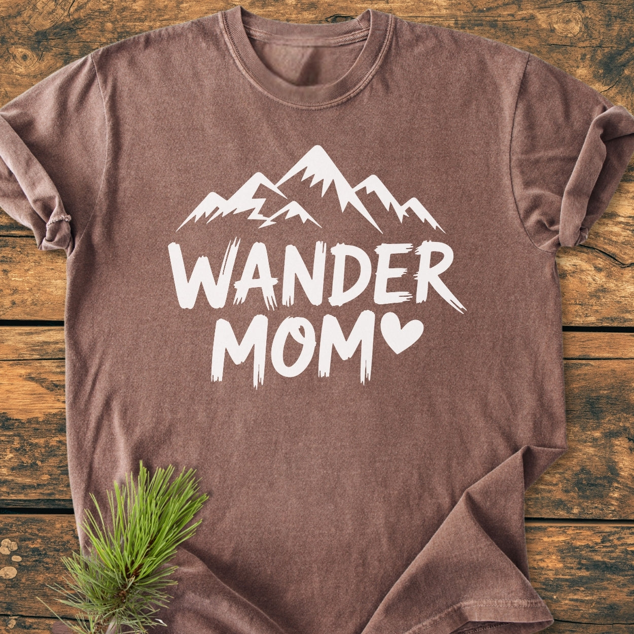Wander Mom - Vintage Wash Tee features stylized mountain peaks and heart-shaped 'O' in 'MOM,' laid flat on a wooden surface with a pine sprig for styling.