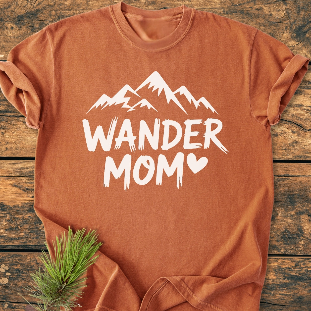 Wander Mom - Vintage Wash Tee: A rust-orange cotton T-shirt featuring a white mountain silhouette and WANDER MOM text, styled with a pine sprig on a wooden surface, conveying an adventure-mama vibe.