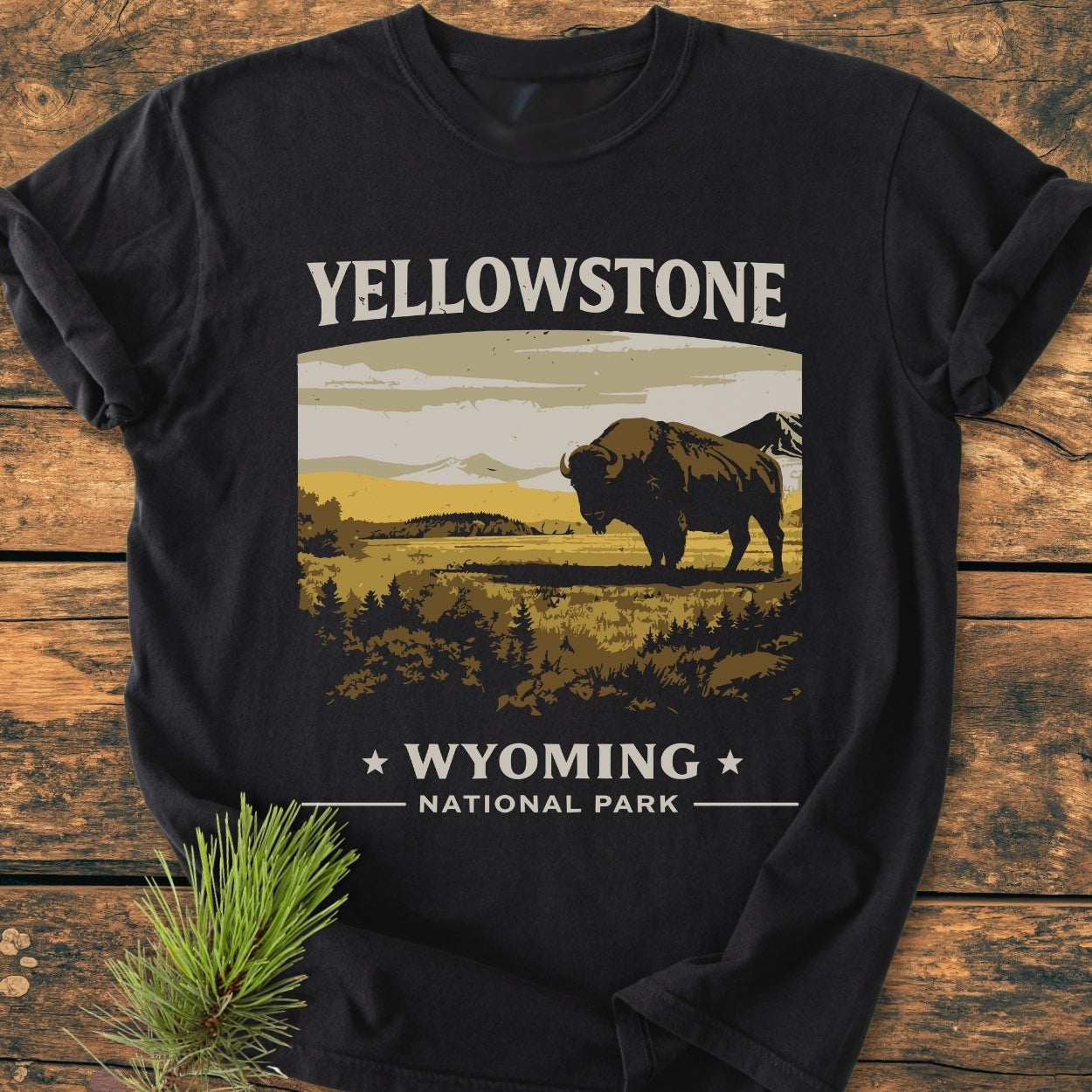 Yellowstone - Vintage Wash Tee featuring a stylized bison graphic with YELLOWSTONE and WYOMING text, laid on a wooden surface with a pine sprig beside it.