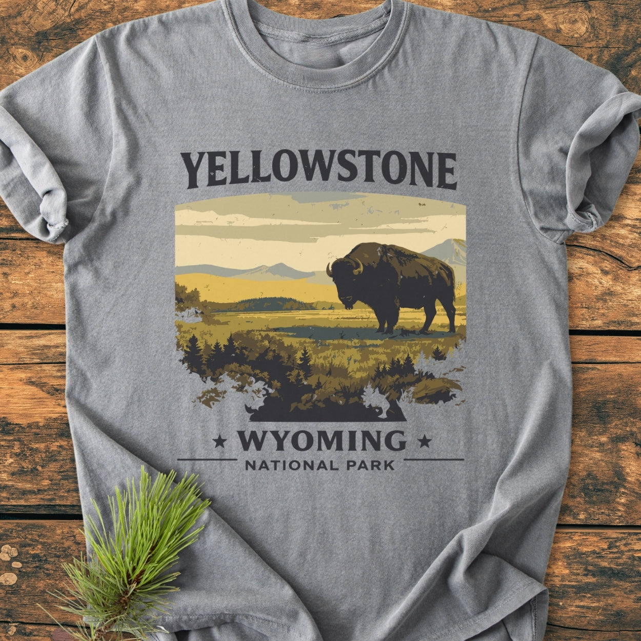 Yellowstone - Vintage Wash Tee featuring a vintage-style print of a bison and mountains, on a heather-gray Comfort Colors 1717 T-shirt, laid on wood with a pine sprig beside it.