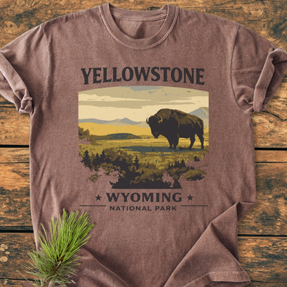 Yellowstone - Vintage Wash Tee featuring a bison graphic on front, laid flat on a wooden surface with a pine bough nearby. Short-sleeve, relaxed fit, made of pre-shrunk, garment-dyed cotton.