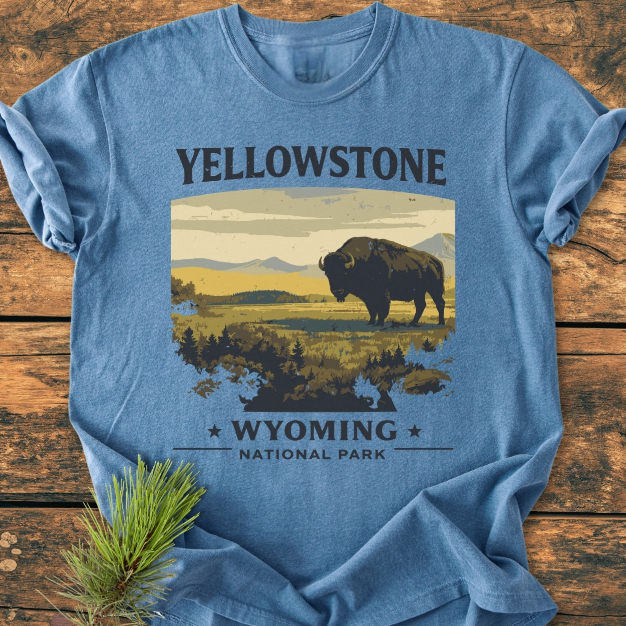 Yellowstone - Vintage Wash Tee featuring a stylized landscape with a bison, mountains, and sky. Laid on wood, it highlights the words “YELLOWSTONE” and “WYOMING NATIONAL PARK,” with a pine sprig beside.
