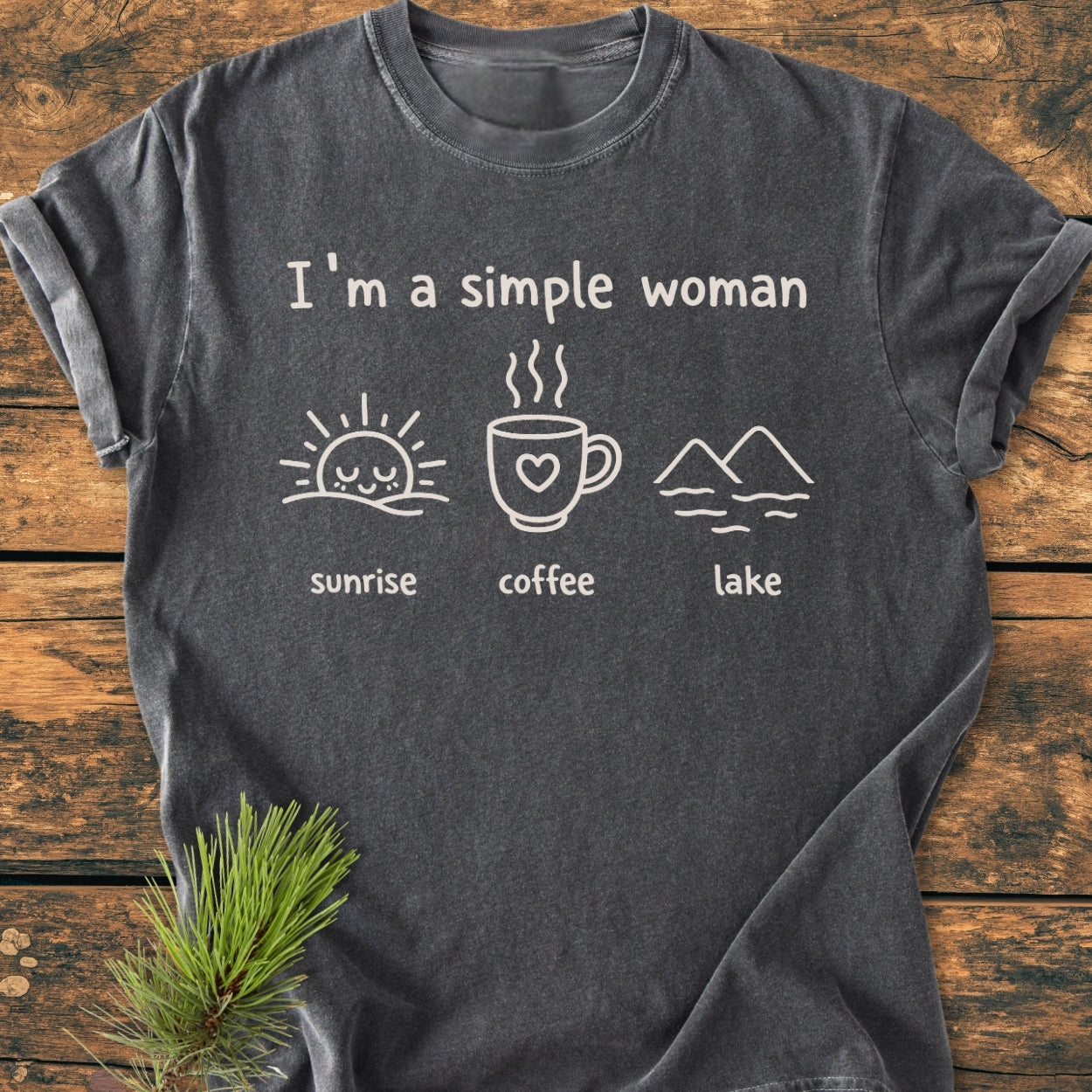 I'm A Simple Woman - Sunrise Coffee Lake - Vintage Wash Tee laid flat, showcasing minimalist line drawings of a sunrise, coffee mug, and lake, styled with a pine sprig on a wooden surface.