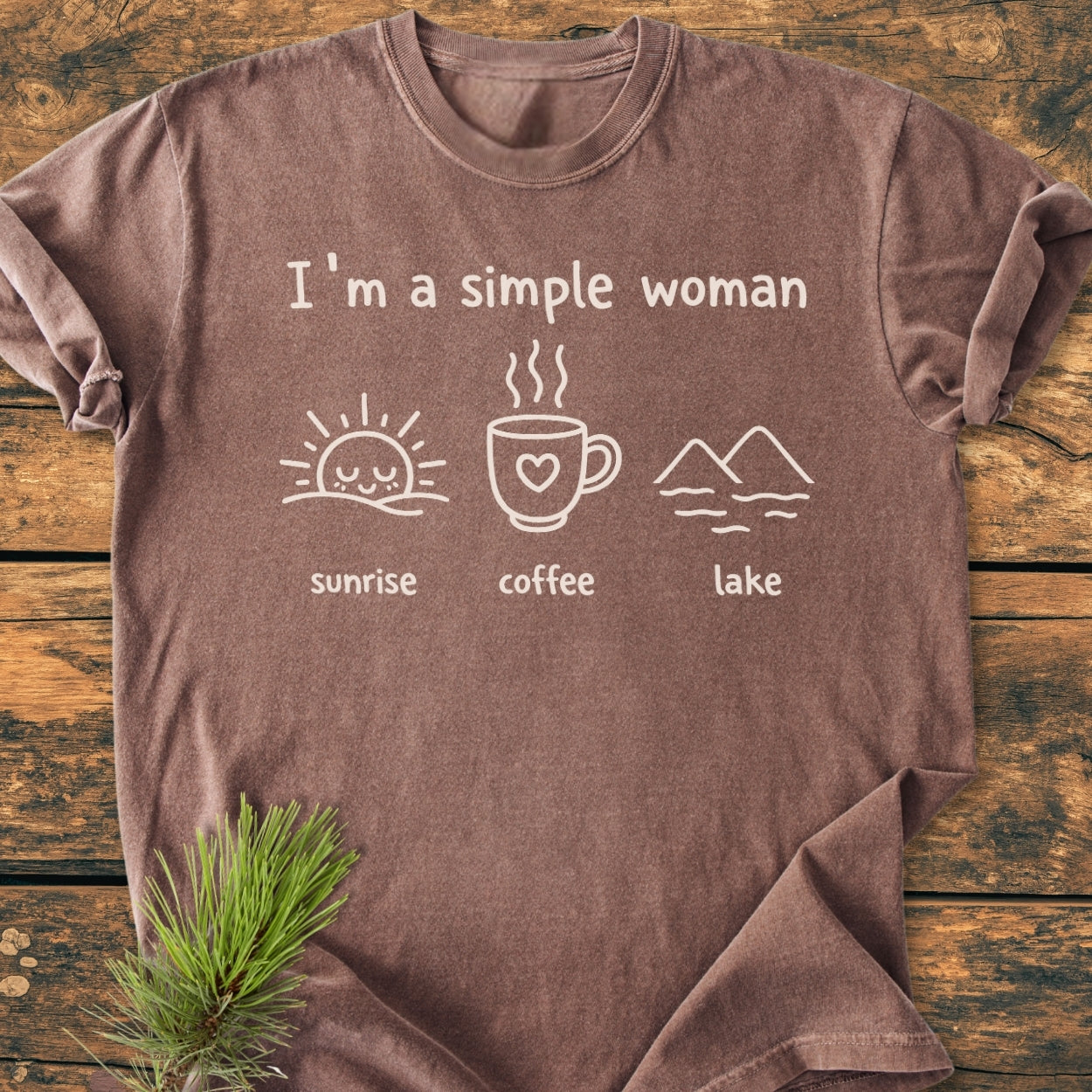 I'm A Simple Woman - Sunrise Coffee Lake - Vintage Wash Tee features a brown tee with white icons of a sunrise, coffee mug, and mountains. Placed on wood, pine sprig accentuates the cozy aesthetic.