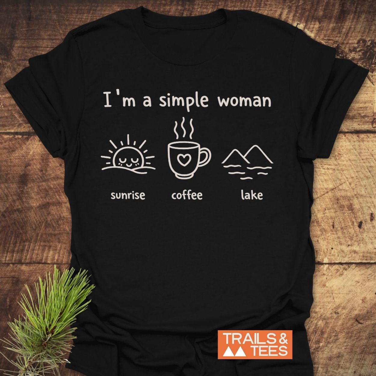 I'm A Simple Woman - Sunrise Coffee Lake T-Shirt displayed on wood, featuring white line-drawn icons of a sunrise, coffee mug, and lake. Includes TRAILS & TEES logo, emphasizing outdoor-inspired comfort.