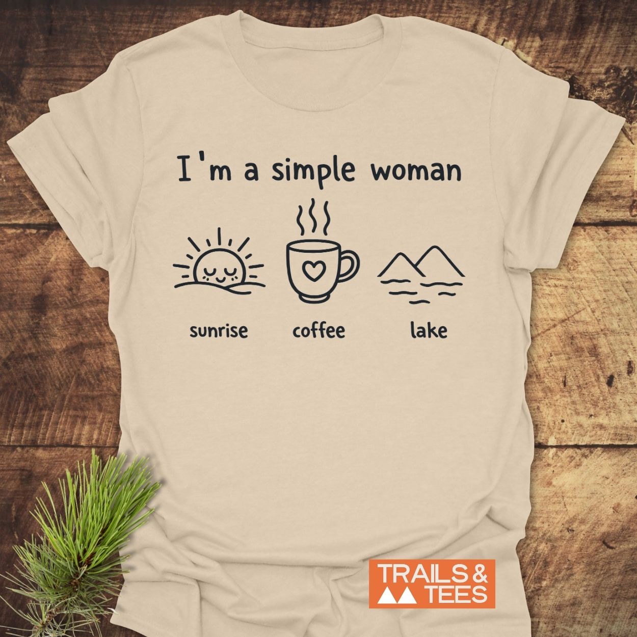 I'm A Simple Woman - Sunrise Coffee Lake T-Shirt, featuring minimalist icons of sunrise, coffee, and lake on light-beige cotton, laid on rustic wood with a pine sprig, Trails & Tees logo visible.