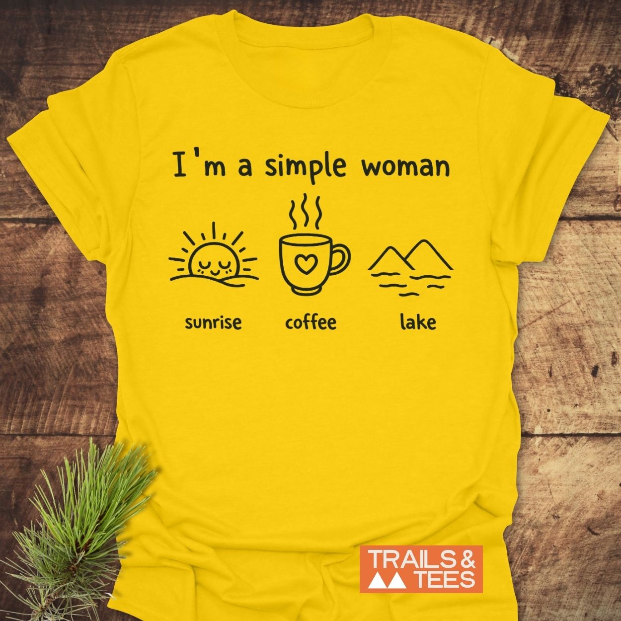 I'm A Simple Woman - Sunrise Coffee Lake T-Shirt featuring minimalist icons of a sunrise, coffee mug, and mountains, laid on wooden surface, with Trails & Tees logo in the corner.