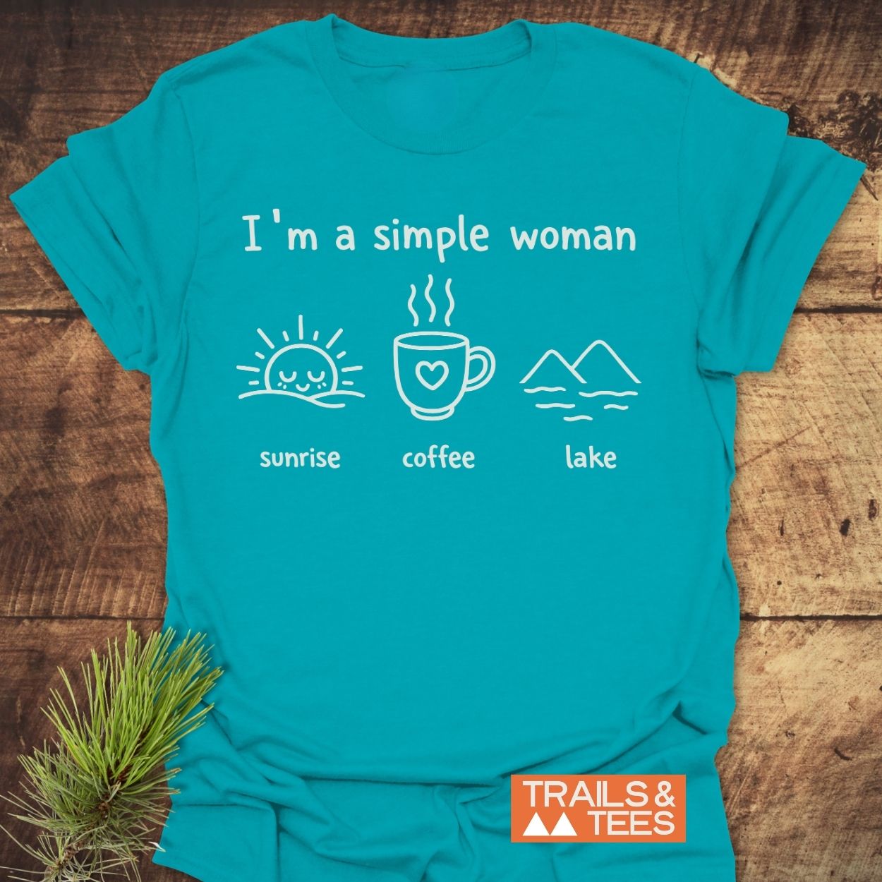 I'm A Simple Woman - Sunrise Coffee Lake T-Shirt flat-lay on wood, featuring white printed text and icons of a sunrise, coffee mug, and lake; TRAILS & TEES in orange at the corner.