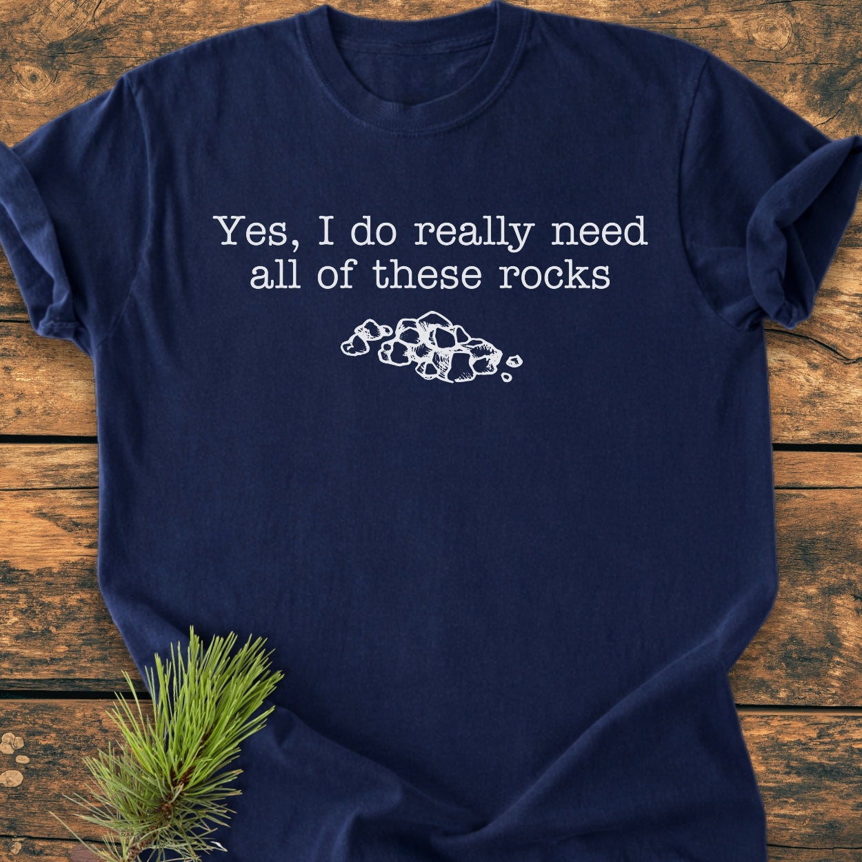 All These Rocks - Vintage Wash Tee features a playful white print on a navy-blue T-shirt, laid out on wood. A pine branch accents this cozy, relaxed-fit Comfort Colors shirt.