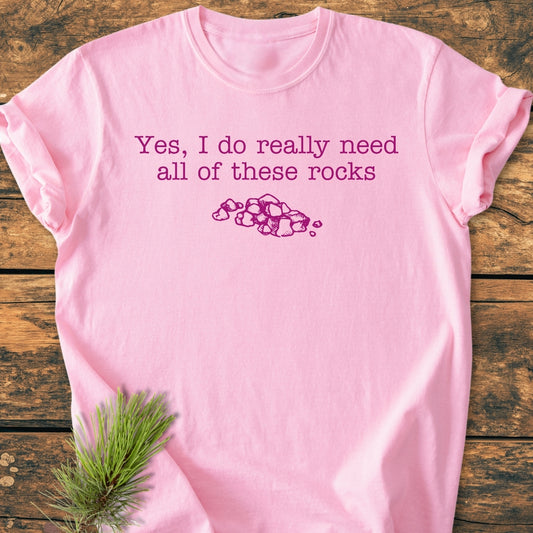 All These Rocks - Vintage Wash Tee features a light pink design with purple text and a rock illustration, laid on a wooden surface, highlighting its relaxed fit and soft-washed fabric.