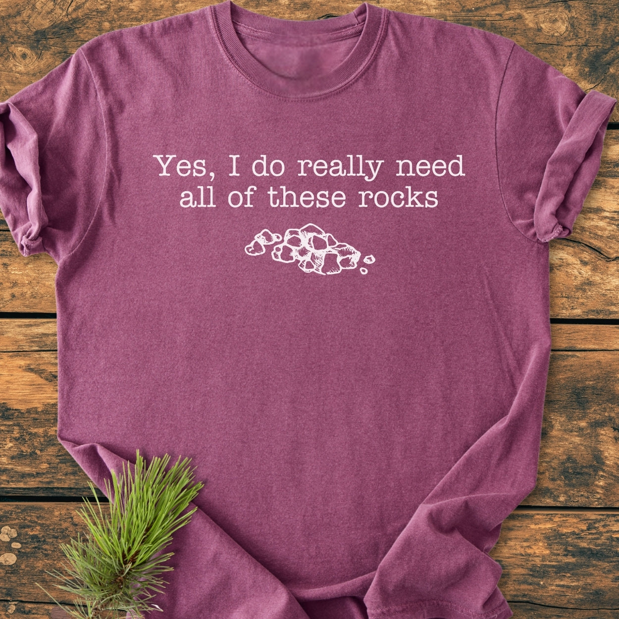 All These Rocks - Vintage Wash Tee featuring a white slogan and stone illustration on a heathered plum T-shirt, laid flat on rustic wood, with a pine sprig accentuating its cozy, relaxed design.