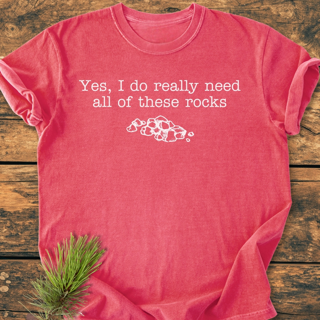 All These Rocks - Vintage Wash Tee, featuring white text and rock line-drawing, is laid on rustic wood with pine needles. Made from soft garment-dyed cotton, it offers a relaxed fit.