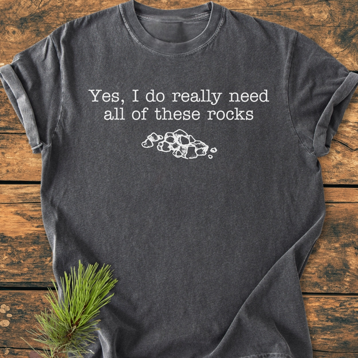 All These Rocks - Vintage Wash Tee in charcoal-gray, featuring a slogan and stone illustration, laid on wood with a pine sprig, highlighting its relaxed fit and comfort-focused design.