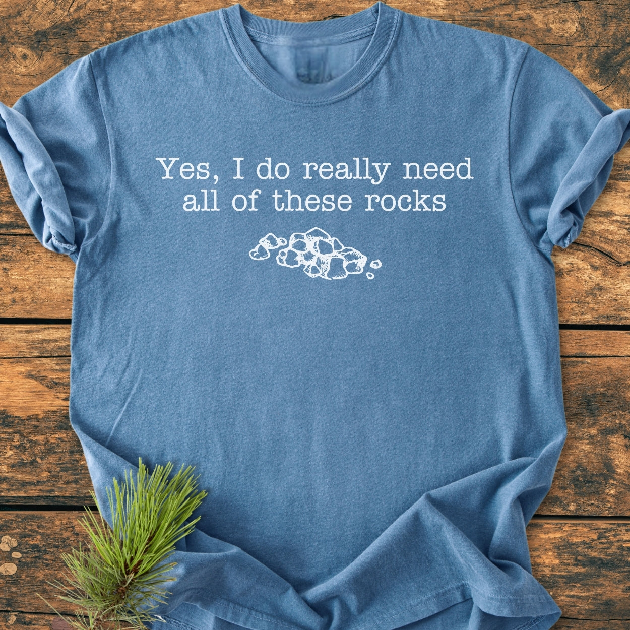 The All These Rocks - Vintage Wash Tee features a heather-blue color with white text and a rock illustration, displayed flat on wood, complemented by a pine sprig, showcasing its relaxed fit.