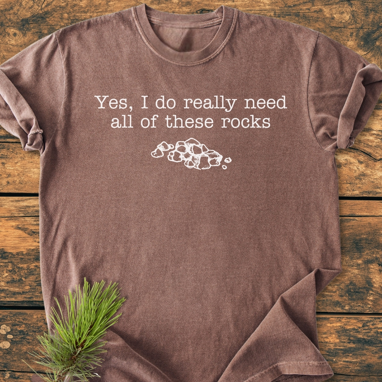 All These Rocks - Vintage Wash Tee: Brown T-shirt with white typewriter-style text, Yes, I do really need all of these rocks, above a rock drawing. Laid on wood with a pine branch nearby.