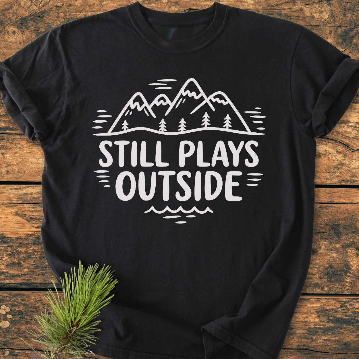 Still Plays Outside - Vintage Wash Tee features a mountain-and-forest graphic with bold lettering on a black t-shirt, laid on wood, highlighting its relaxed fit and soft-washed, garment-dyed fabric.