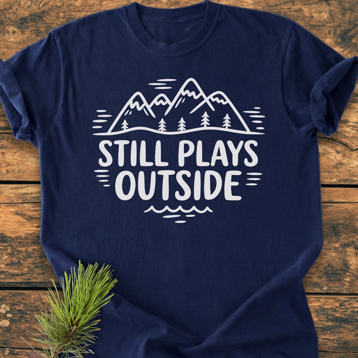 Still Plays Outside - Vintage Wash Tee featuring white mountain and pine tree graphic on navy, laid on wood with pine sprig; relaxed fit, durable, garment-dyed Comfort Colors 1717 shirt.