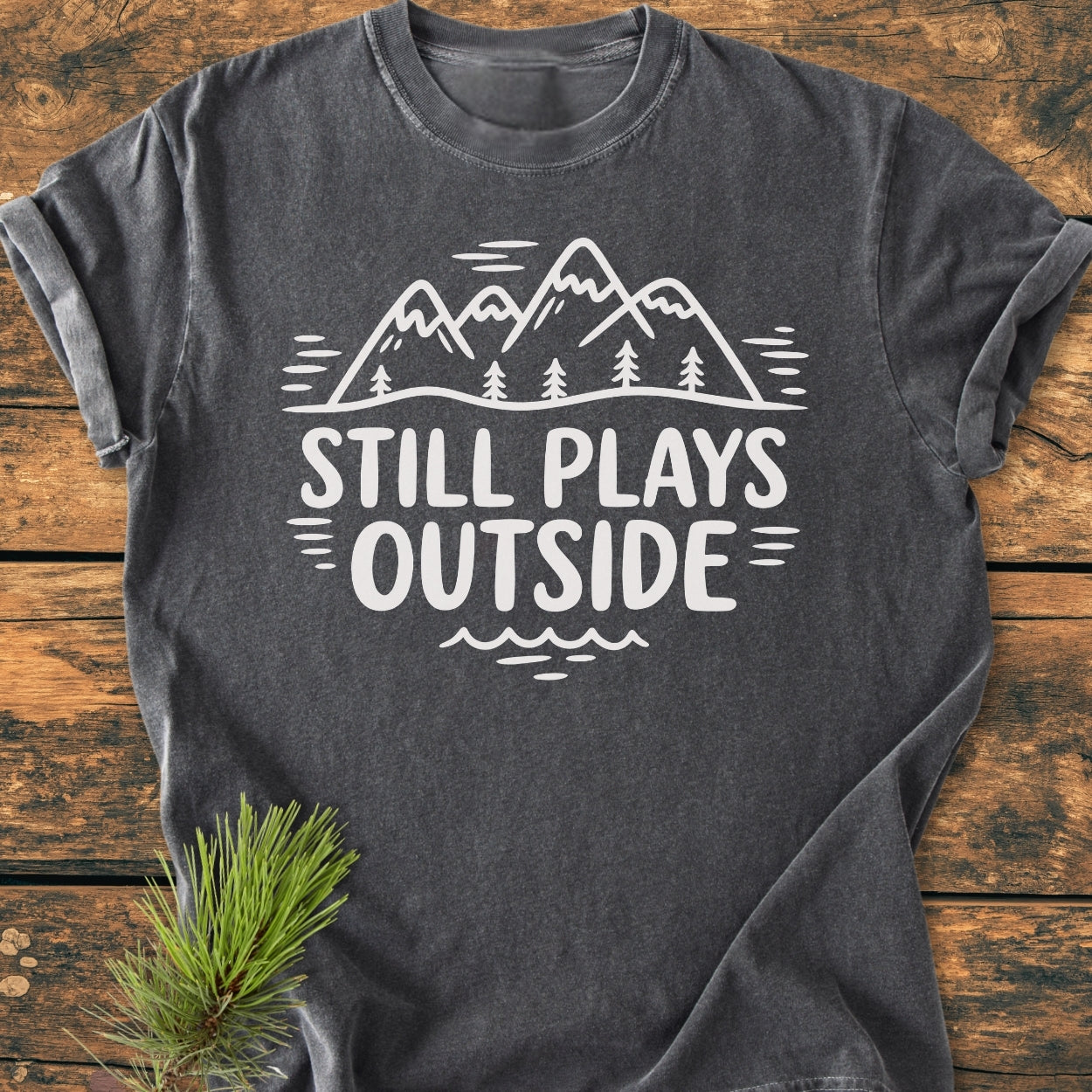 Charcoal-gray Still Plays Outside - Vintage Wash Tee, showcasing a mountain-and-trees graphic. Set on a rustic wooden background with a pine sprig accent, highlighting its cozy, relaxed fit and durable design.