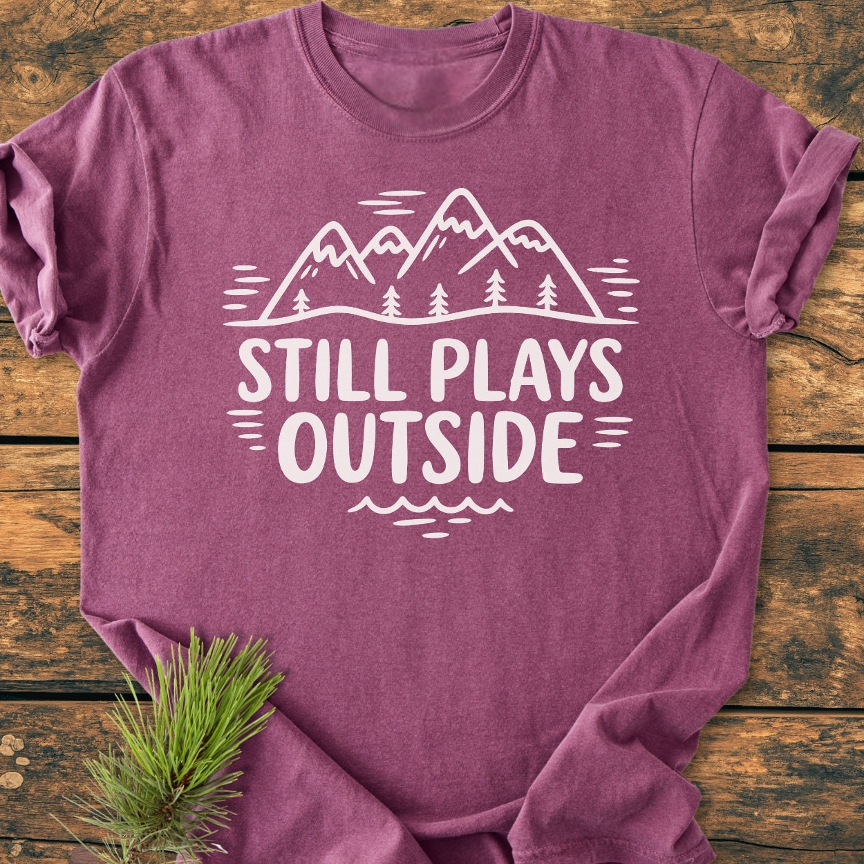 Still Plays Outside - Vintage Wash Tee featuring a mountain-and-trees graphic and slogan on a mauve crew-neck, laid flat on wood, with a pine sprig beside.