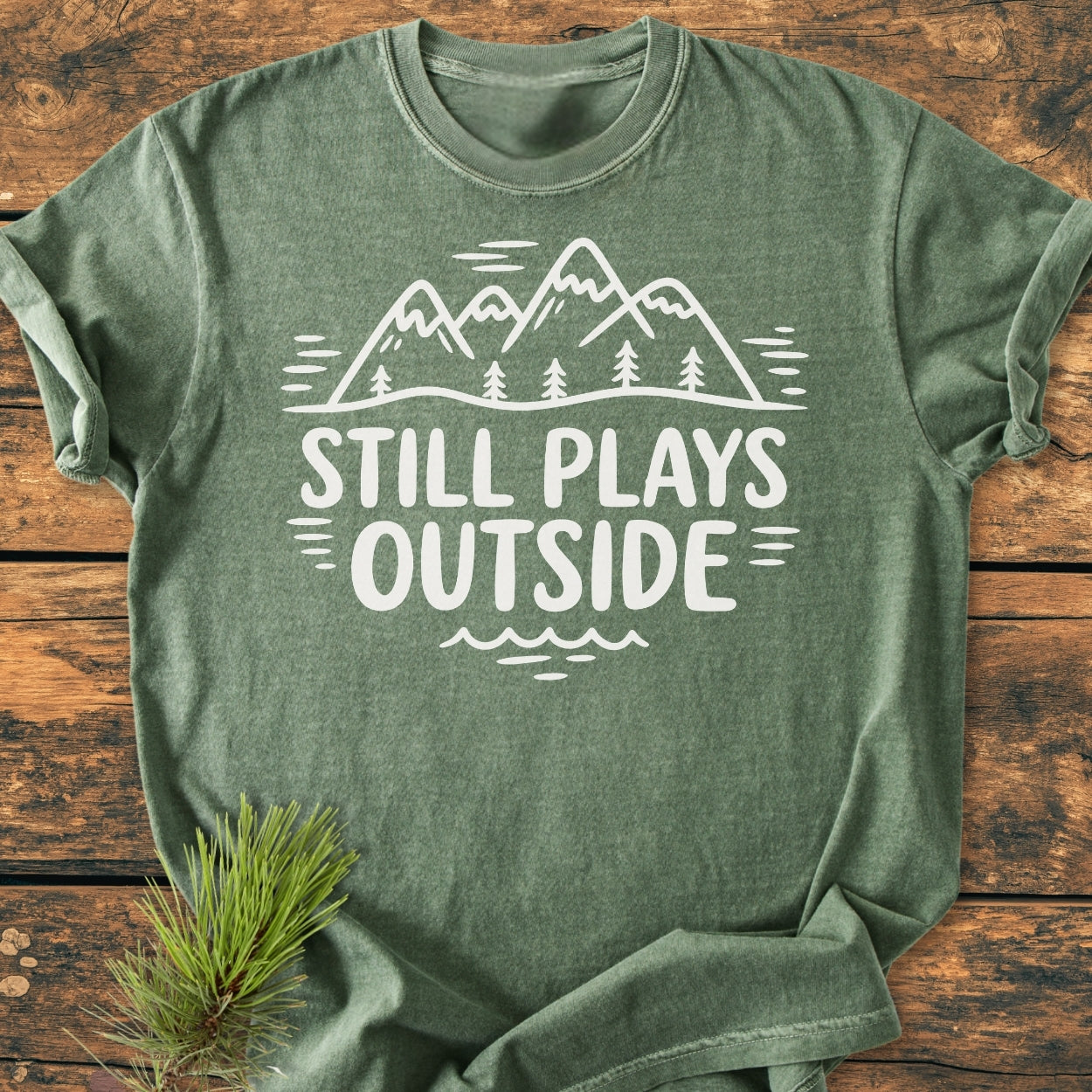 Heather-green Still Plays Outside - Vintage Wash Tee with a white mountain-and-pine-tree graphic and slogan, laid on wood surface with pine needles. Made from soft, durable, pre-shrunk ring-spun cotton.