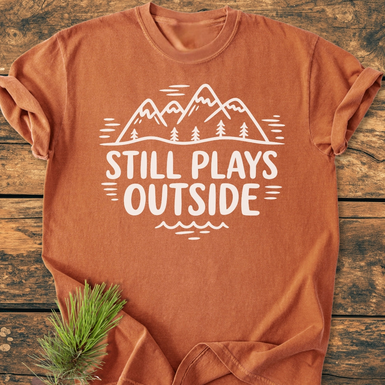 Still Plays Outside - Vintage Wash Tee, featuring a mountain-and-tree design with slogan, laid flat on wood. Soft, rust-orange cotton enhances its relaxed, cozy appeal, complemented by a rustic pine branch accent.