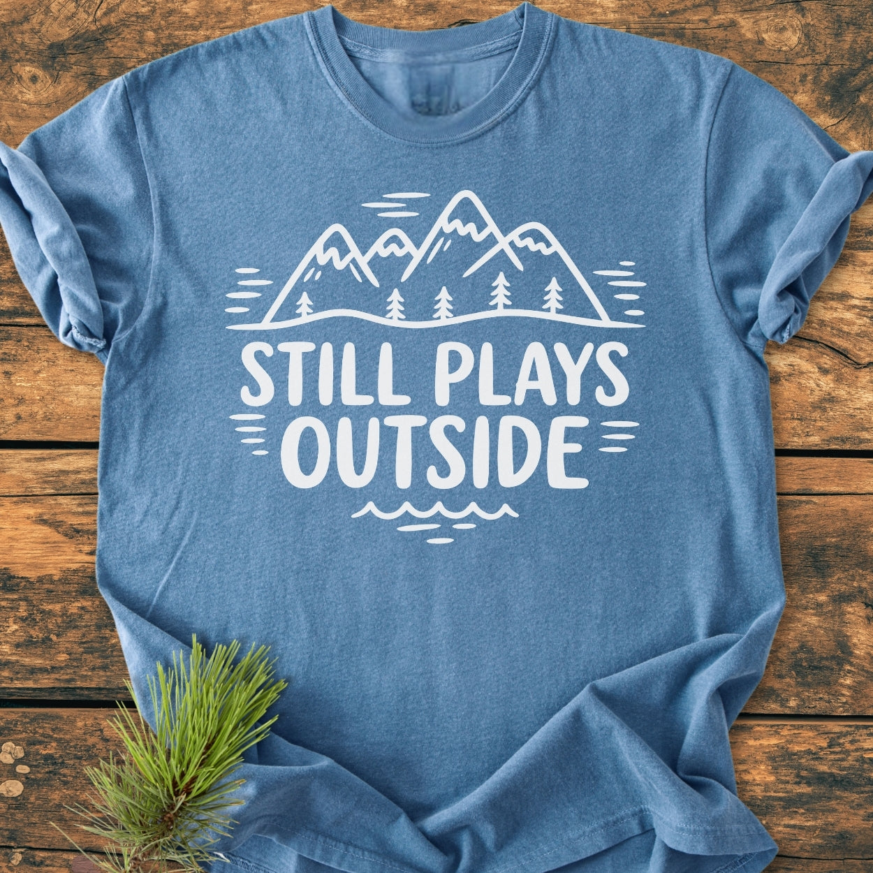 Still Plays Outside - Vintage Wash Tee: Heather-blue cotton, featuring white mountain peaks and pine trees graphic with bold text. Laid flat on wood, includes pine needle accent, highlighting its outdoor-inspired design.