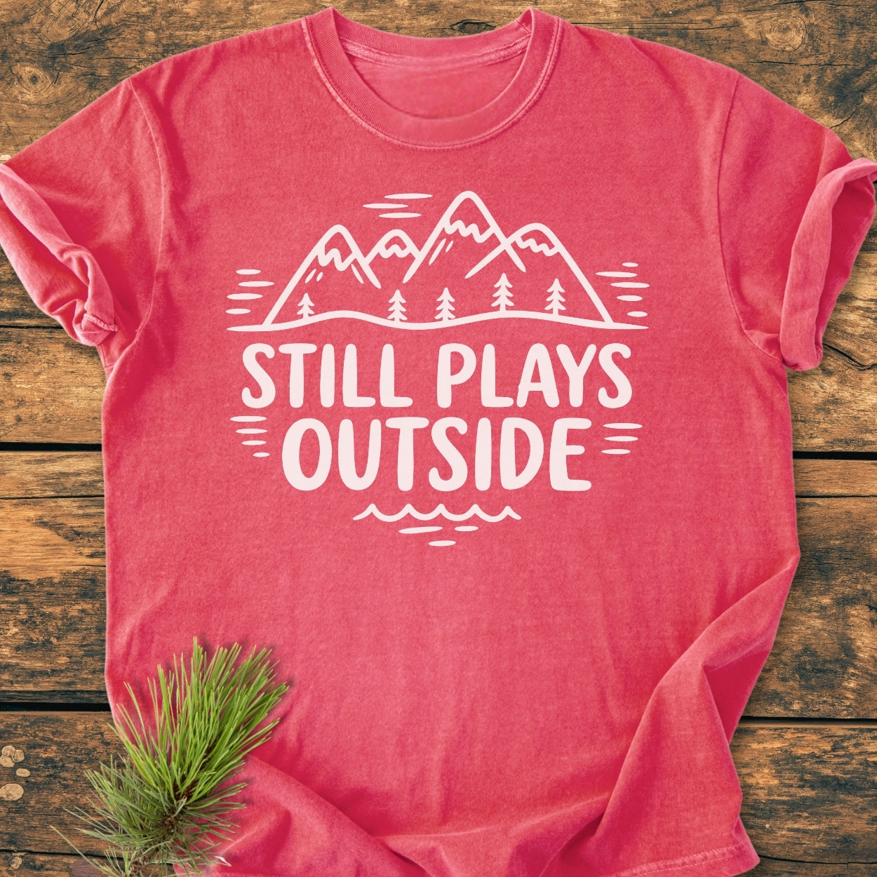 Still Plays Outside - Vintage Wash Tee features a mountain and pine tree line-art design on a rustic wooden surface, showcasing its relaxed fit and durable stitching for everyday wear.