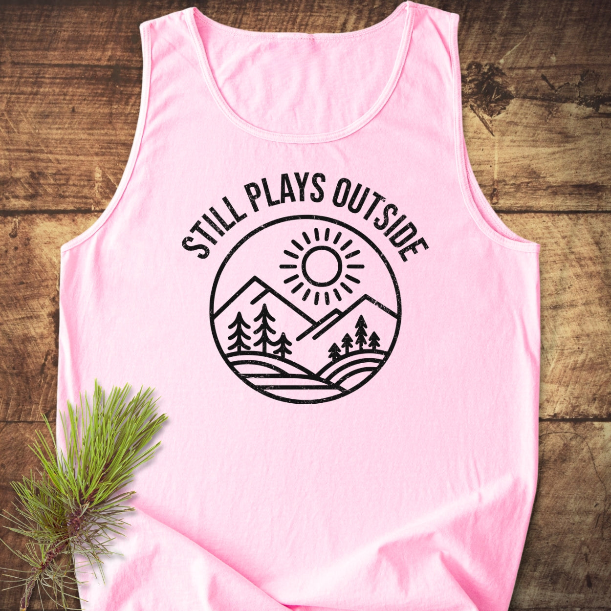 Light-pink Still Plays Outside tank top with a black circular graphic of mountains, trees, and sun, laid on wood with a pine branch. Made from 100% US cotton, offering a relaxed fit.