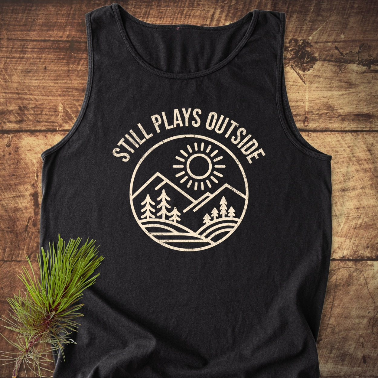 Still Plays Outside Tank Top on rustic wood, featuring a cream mountain and sun graphic. Unisex, relaxed fit, made from sustainable, 100% ring-spun US cotton with eco-friendly dyes.