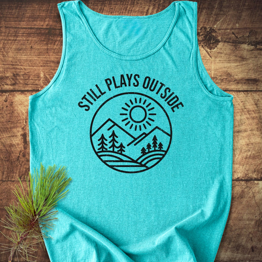 Still Plays Outside Tank Top featuring a black badge-style graphic of mountains, trees, and a sun, laid flat on a wooden surface with a pine branch for a natural touch.