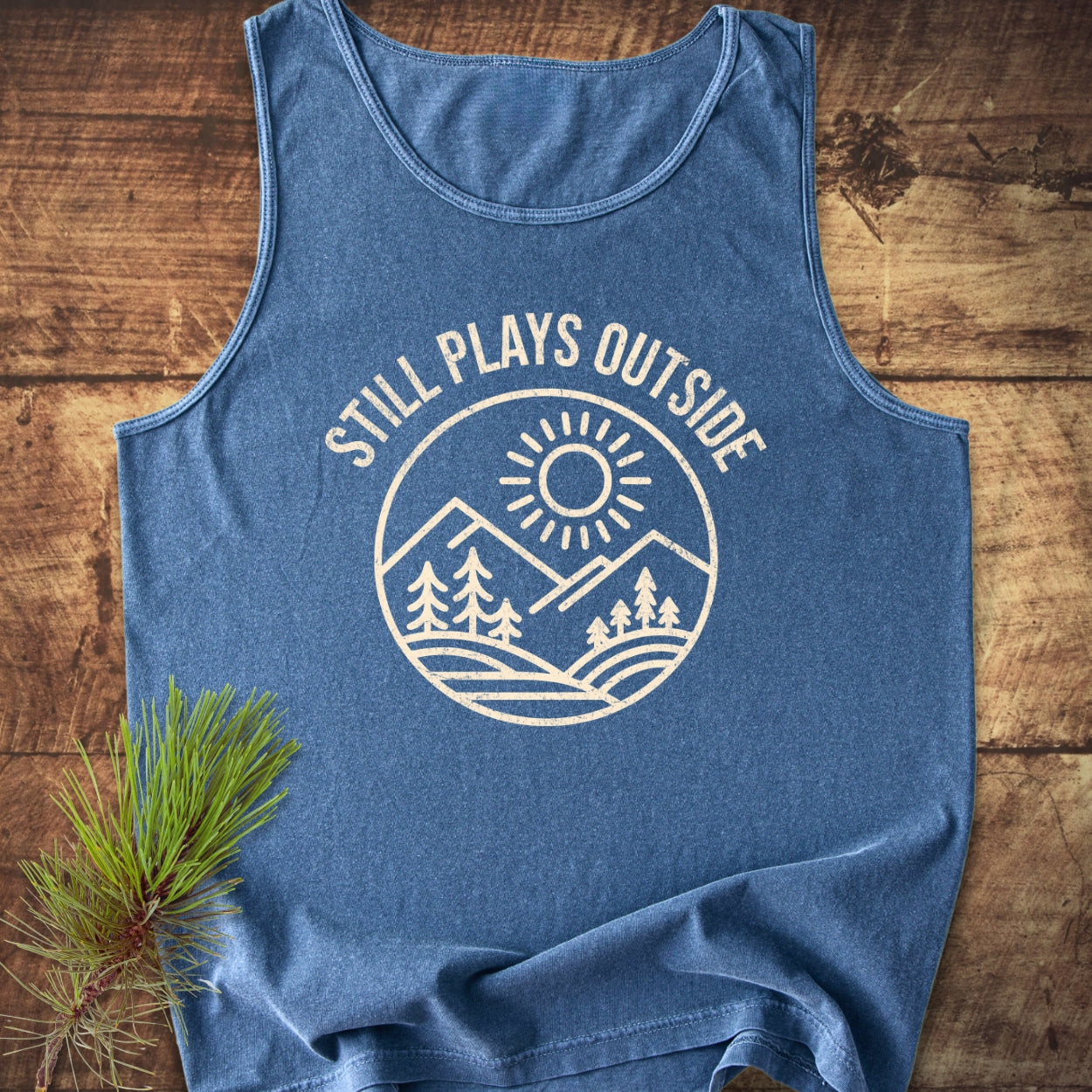Still Plays Outside Tank Top with a round badge design featuring a sun over mountains and trees; laid on rustic wood, complemented by a pine branch for a nature-inspired touch.