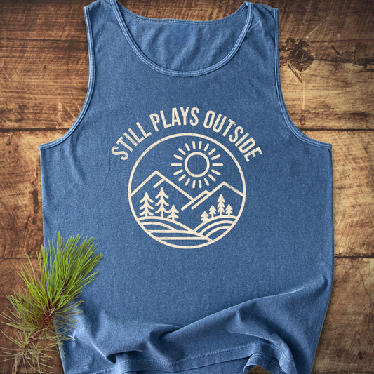Still Plays Outside Tank Top with a round badge design featuring a sun over mountains and trees; laid on rustic wood, complemented by a pine branch for a nature-inspired touch.