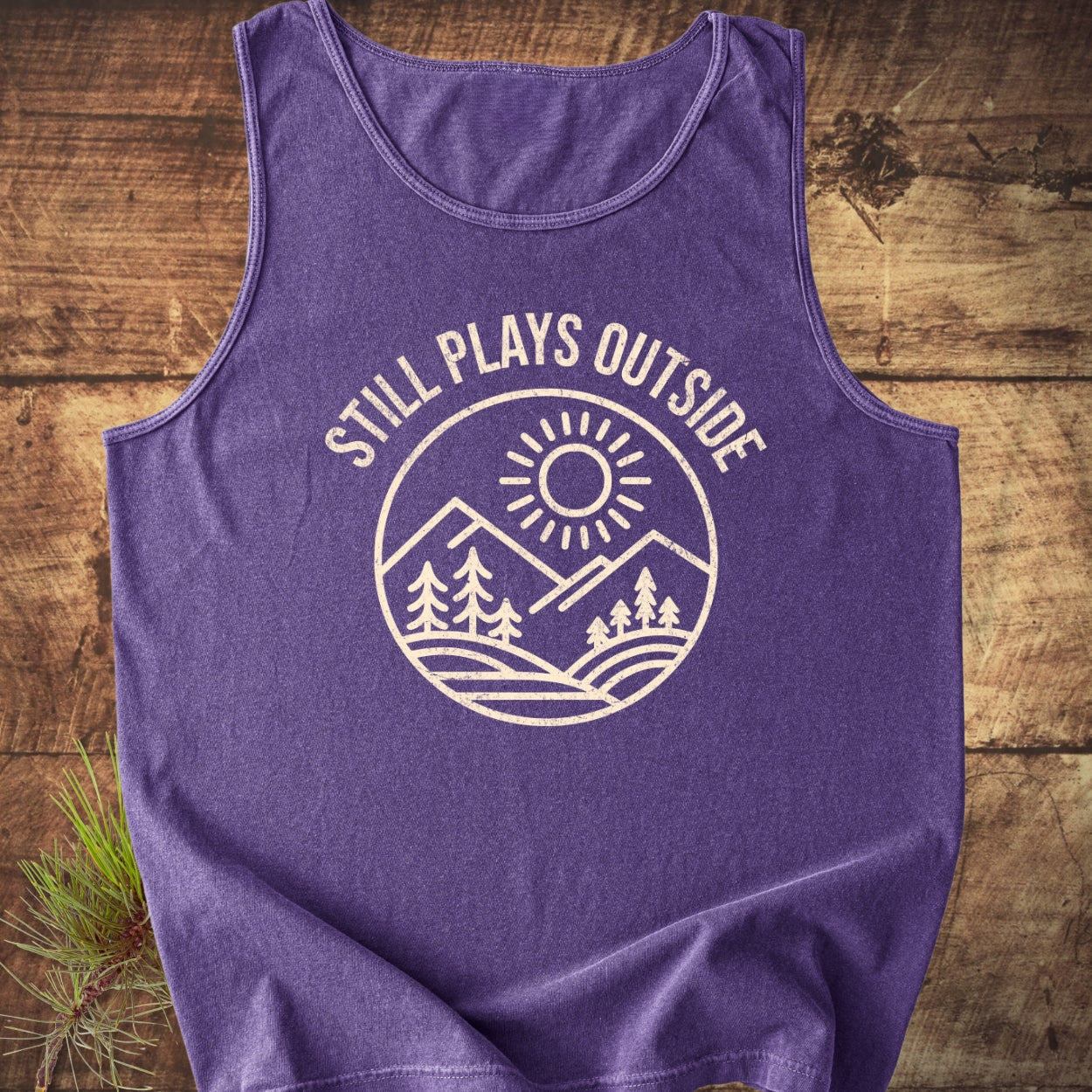 Still Plays Outside Tank Top with a circular graphic of mountains and trees, laid on wood. Features a relaxed fit, made from sustainable US cotton with eco-friendly dyes, showcasing rustic outdoor vibes.