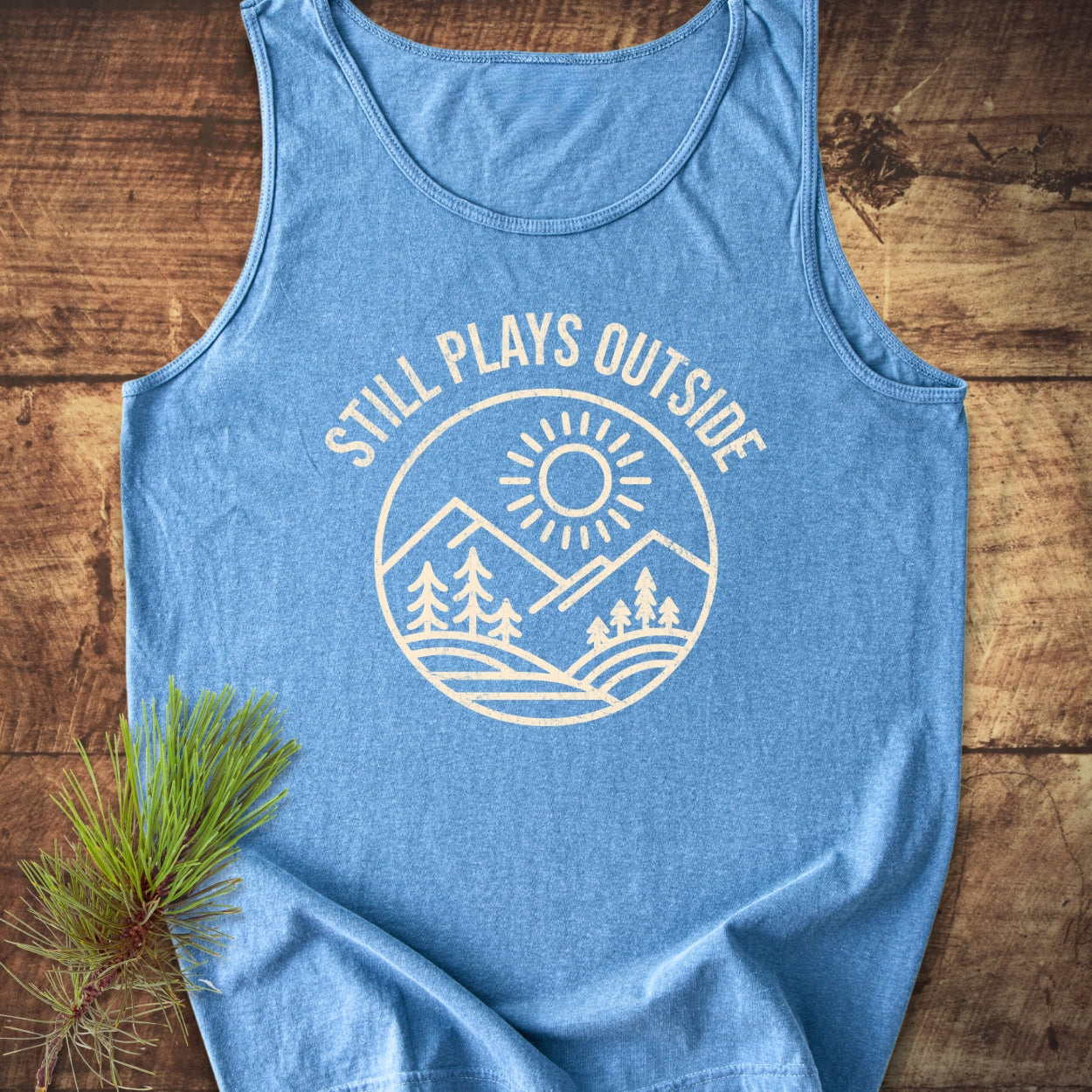 Still Plays Outside Tank Top: Heather-blue tank featuring cream circular mountain and sun design with STILL PLAYS OUTSIDE text, laid on wood with pine branch, highlighting comfort and sustainability.