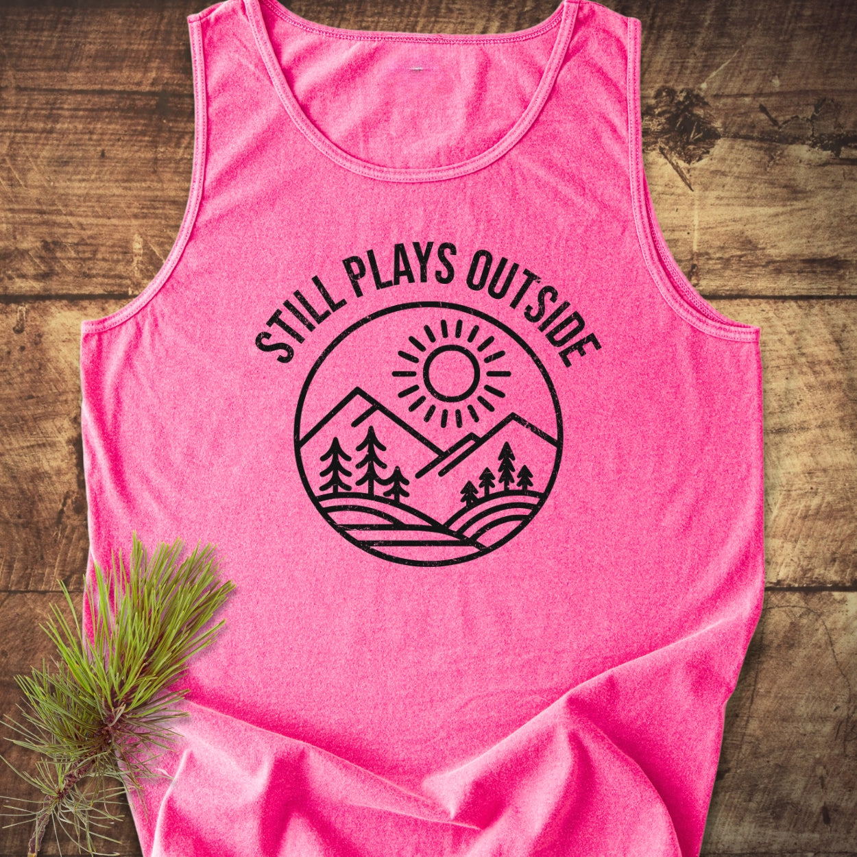 Still Plays Outside Tank Top, showcasing a circular line-art graphic of nature themes, laid flat on a wooden surface with pine needles, crafted from sustainable, comfortable 100% ring-spun US cotton.
